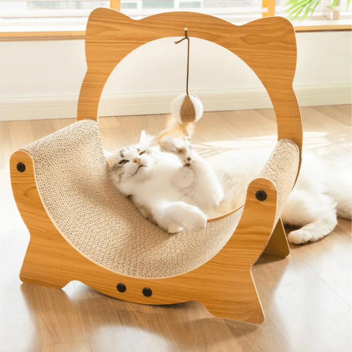 Na imagem, há um gato persa branco e cinza, de aparência fofa e pelagem densa, brincando com um brinquedo suspenso. Ele está deitado de costas em uma peça de mobiliário para gatos que tem uma estrutura em forma de cabeça de gato, feita de madeira clara com acabamento liso. A área central onde o gato está deitado é feita de papelão, que é adequado para que os gatos arranhem e estiquem suas garras.