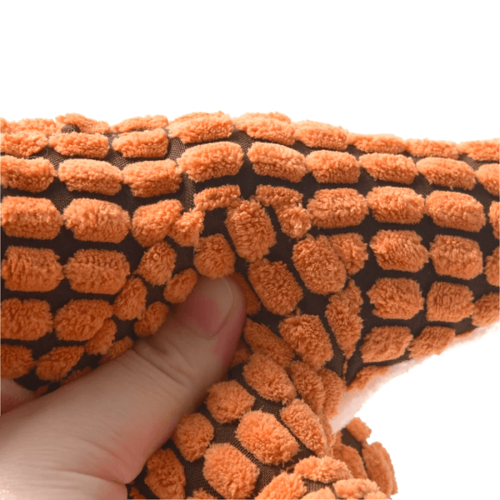 Close-up da textura de um tecido de brinquedo para cachorro, mostrando uma mão humana apertando o material. O tecido tem um padrão de pequenas protuberâncias laranja sobre um fundo marrom escuro, demonstrando a superfície macia e emborrachada que é agradável ao toque e ideal para cães mastigarem.