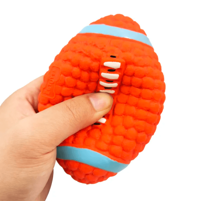 Mão humana segurando um brinquedo para cachorro em formato de bola de futebol americano. A bola é de borracha com textura saliente, na cor laranja vibrante e com listras azuis claras e costuras brancas decorativas. A imagem foca no brinquedo e na mão, proporcionando uma boa noção do tamanho e da escala do objeto.