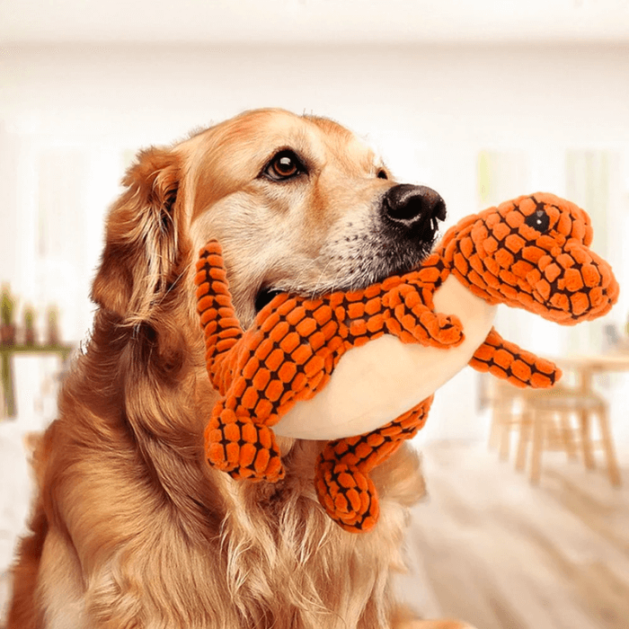 Um Golden Retriever com pelo dourado segura gentilmente um brinquedo de pelúcia em forma de dinossauro Tiranossauro Rex laranja e creme. O cão olha para a esquerda com uma expressão atenta e carinhosa, enquanto o brinquedo parece ser macio e confortável em sua boca, sugerindo uma cena de brincadeira agradável e tranquila em um ambiente interno bem-iluminado.